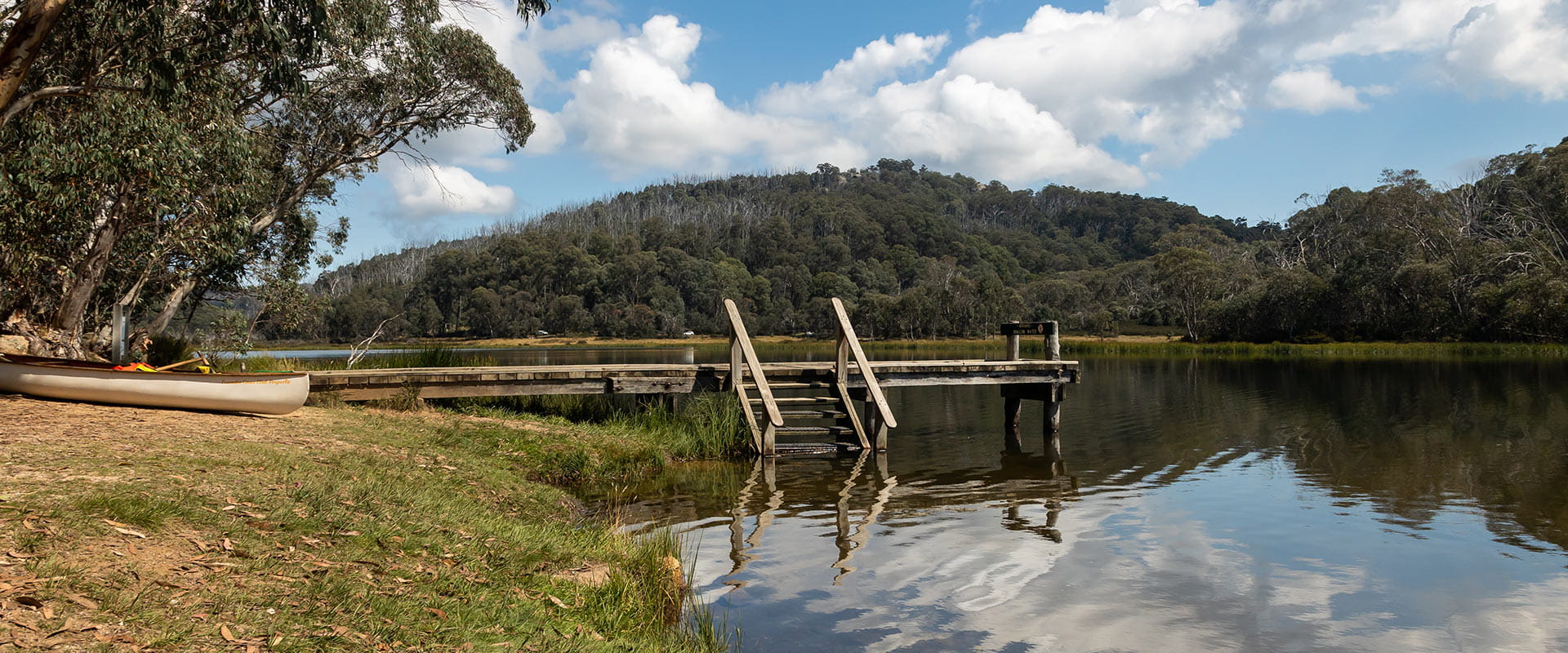 A small wooden jetty extends from the lake shore, next to a canoe and in front of a hill below a blue sky filled fluffy white clouds.