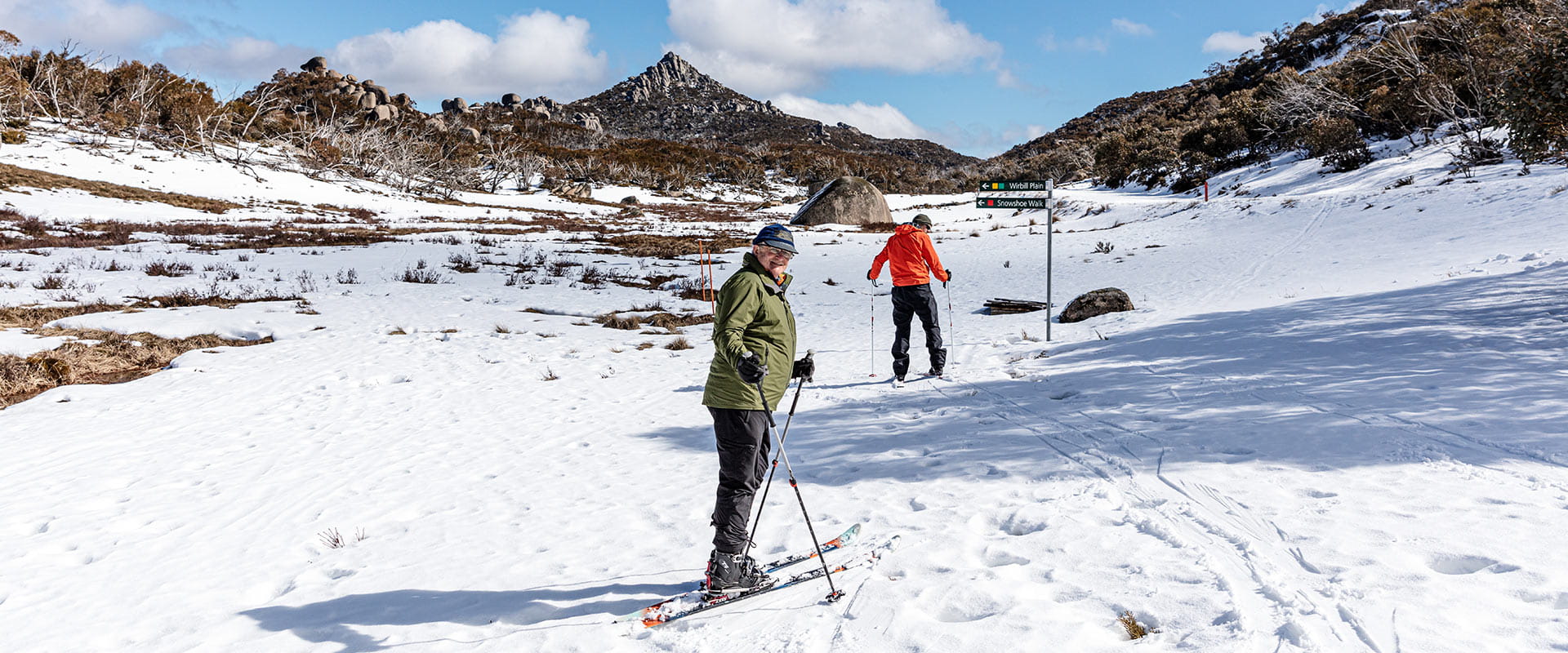An older man stands for the camera while cross country skiing infront of a mountain peak.