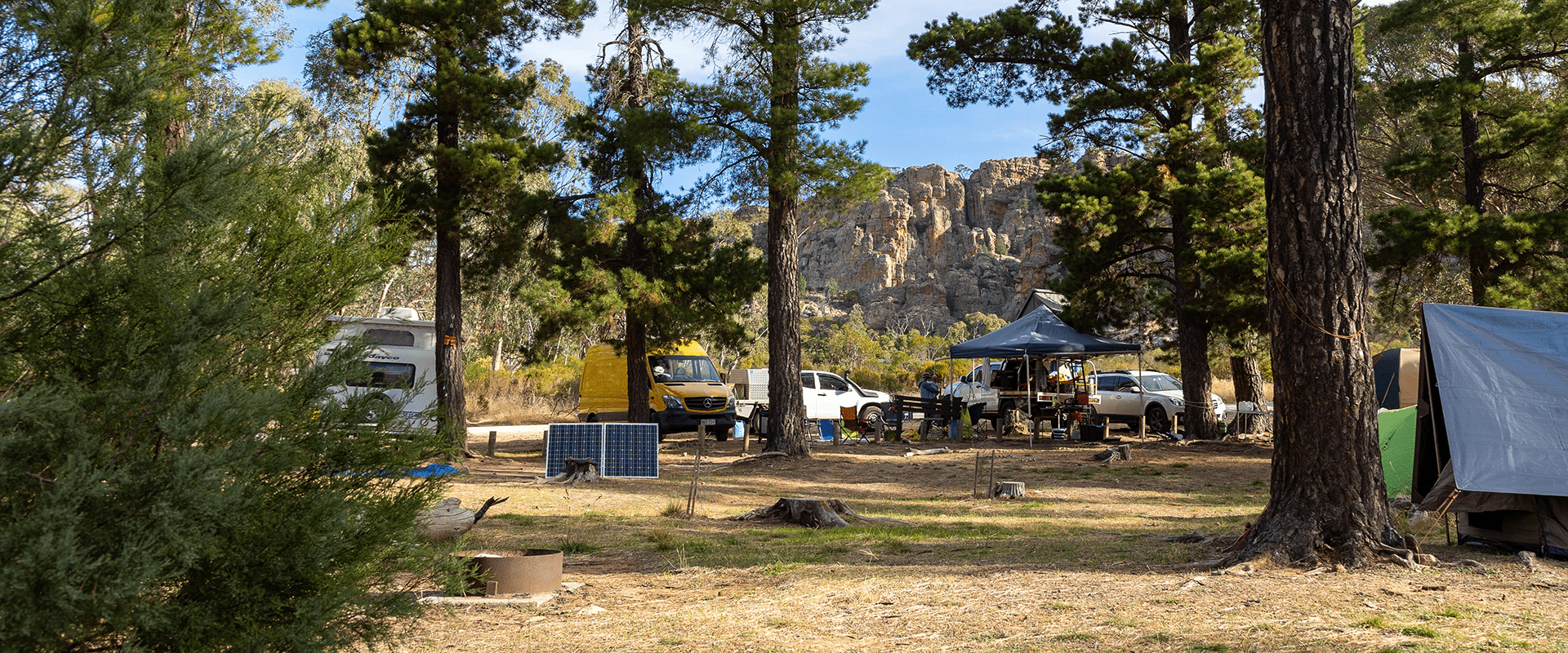 A camp site with tall pine trees with rock climbing boulders in the back ground, there are 4 wheel drives, tents and caravans camping