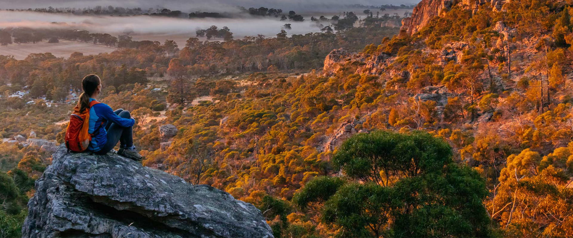 A woman sits high up on a hill on top of a rock overlooking the park and enjoying the sunset which falls on the mountain side