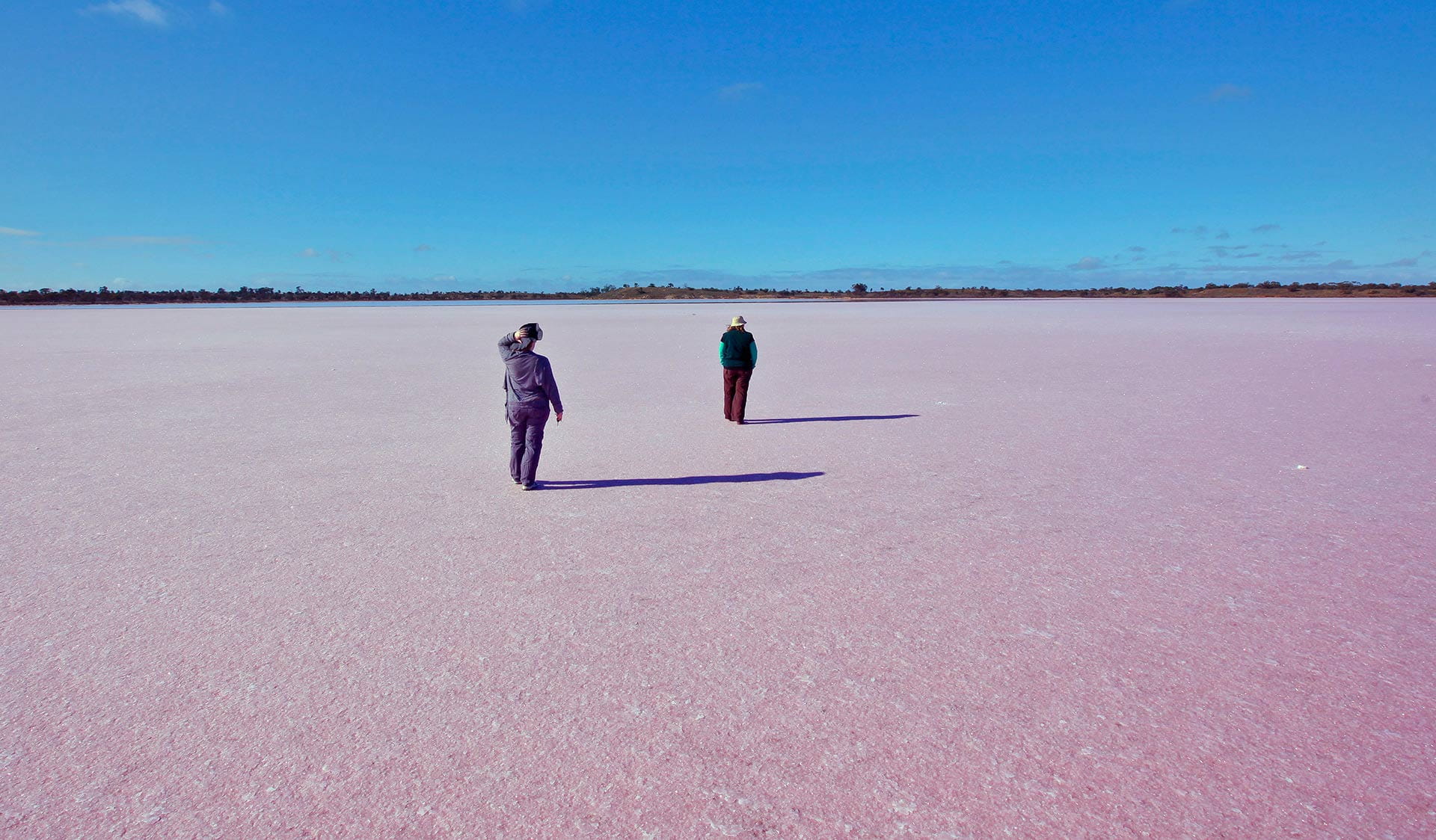 Two people walk across a dry pink salt lake.