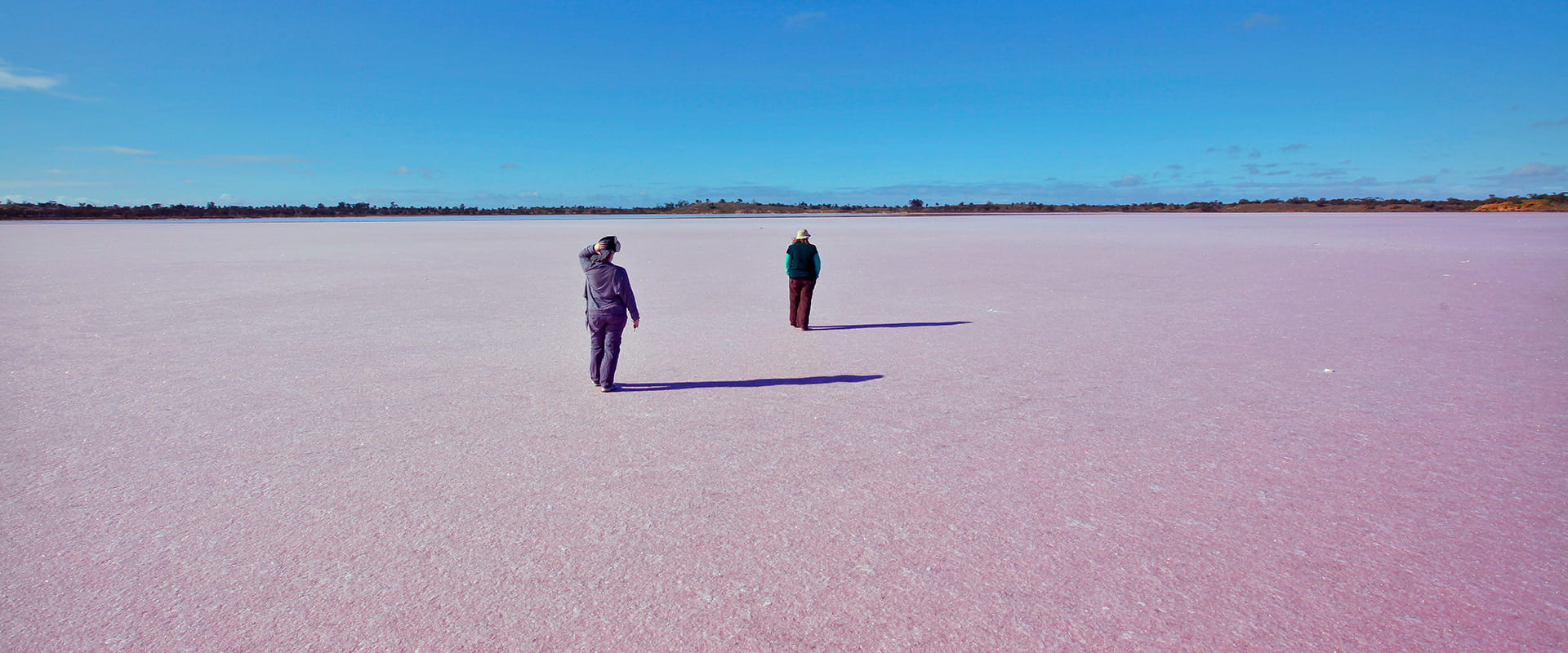 Two people walk across a dry pink salt lake.