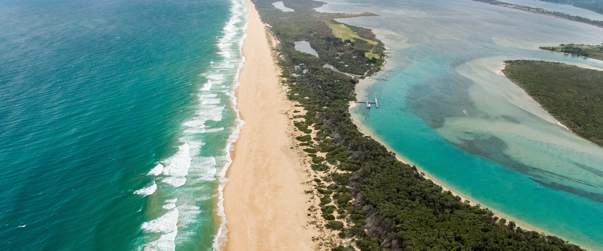 An aerial view that looks down at a secluded stretch of coastline with dunes and coastal shrubbery that seperate the beach from an expansive coastal river system.