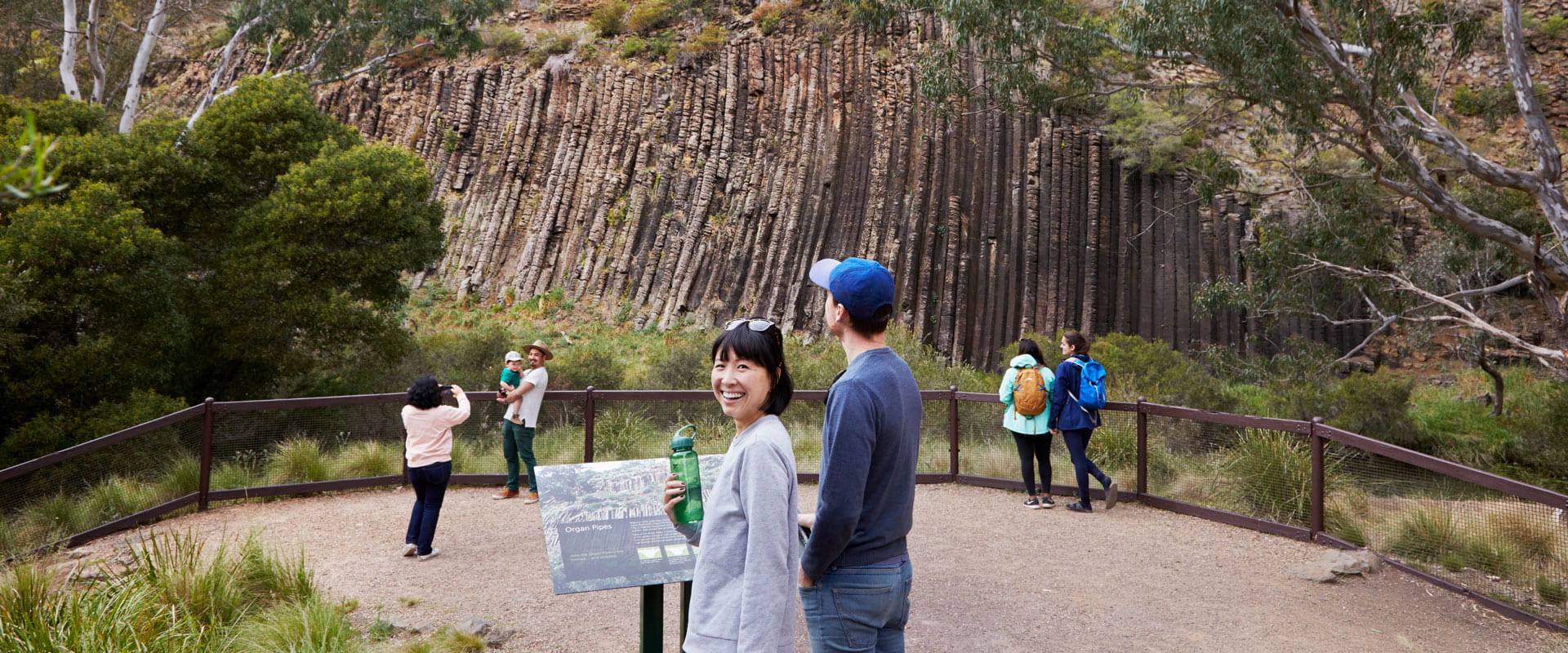 A woman holding a drink bottle stands next to a man admiring the large stone columns reaching up a rocky cliff face as a young family takes a photo in front of the natural structure against a barrier in the background.