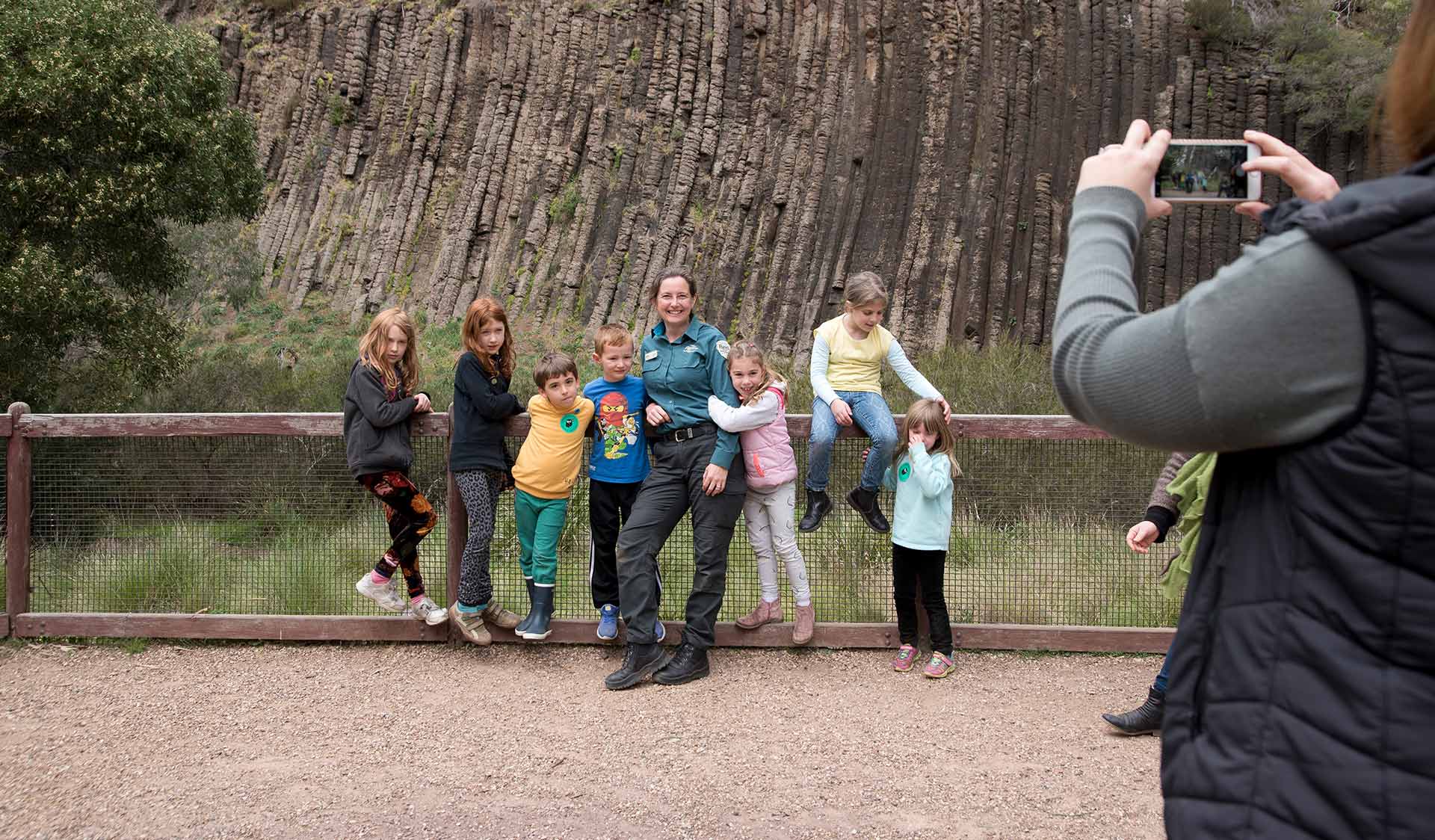 A Parks Victoria Ranger poses for a photograph with 7 young children in the Junior Rangers Program in front of the rock formation at Organ Pipes National Park.