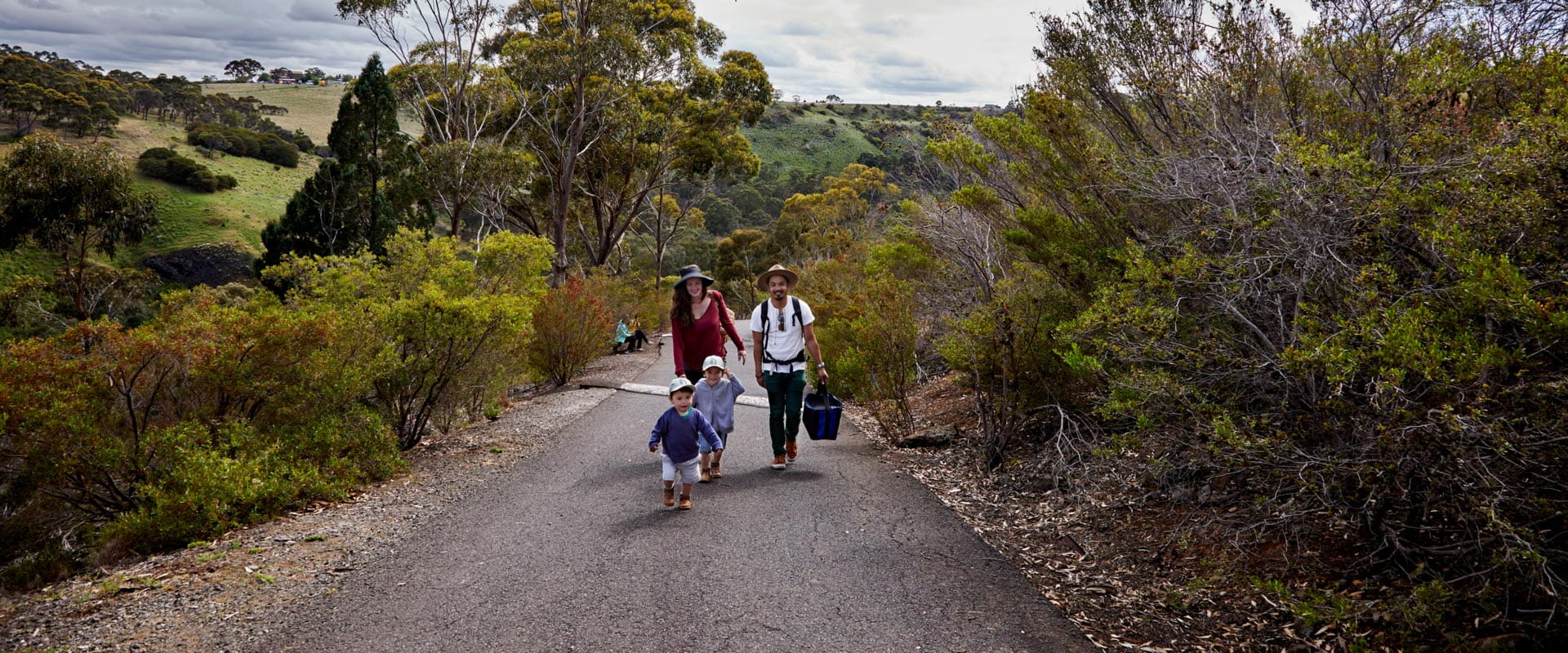 A family with two young children walk up a road path bordered by shrubs, with trees and rolling hills behind them.