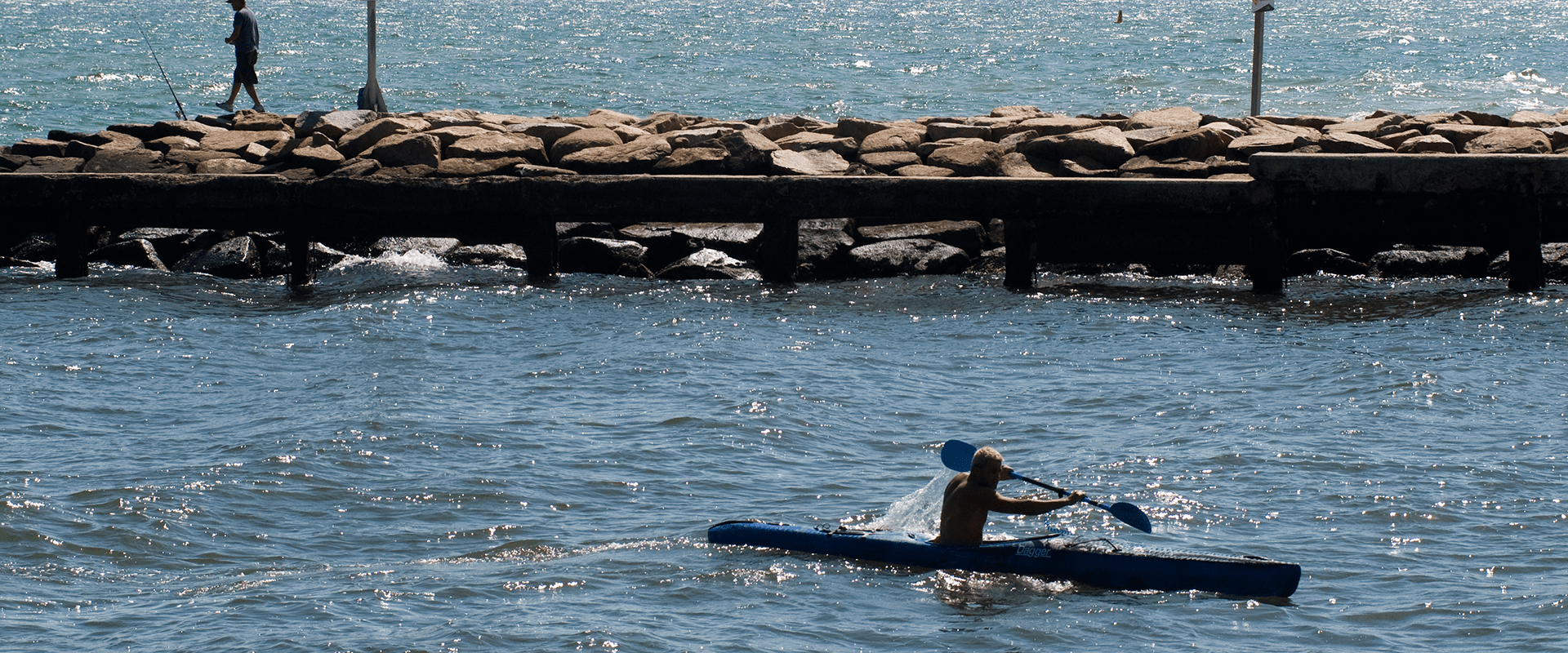 A sunny day at the mouth of the river with the bay in the background. A person is enjoying time in a kayak and another person is fishing off the breakwater.
