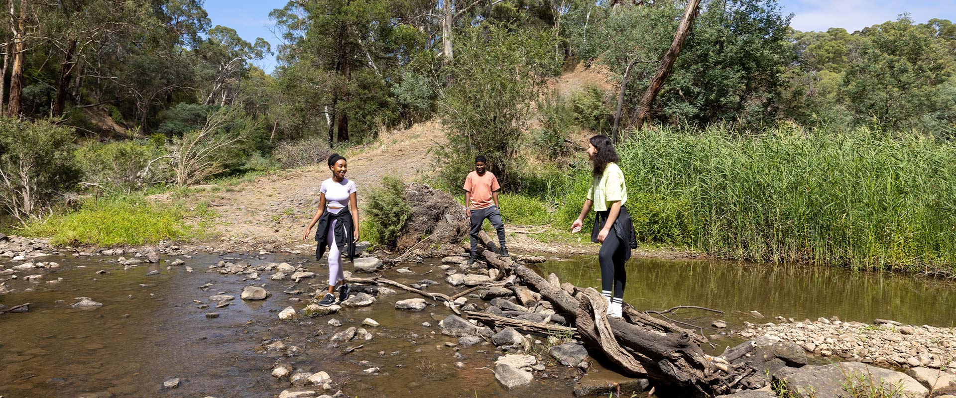 Three people from CALD backgrounds cross a shallow river by stepping across stones
