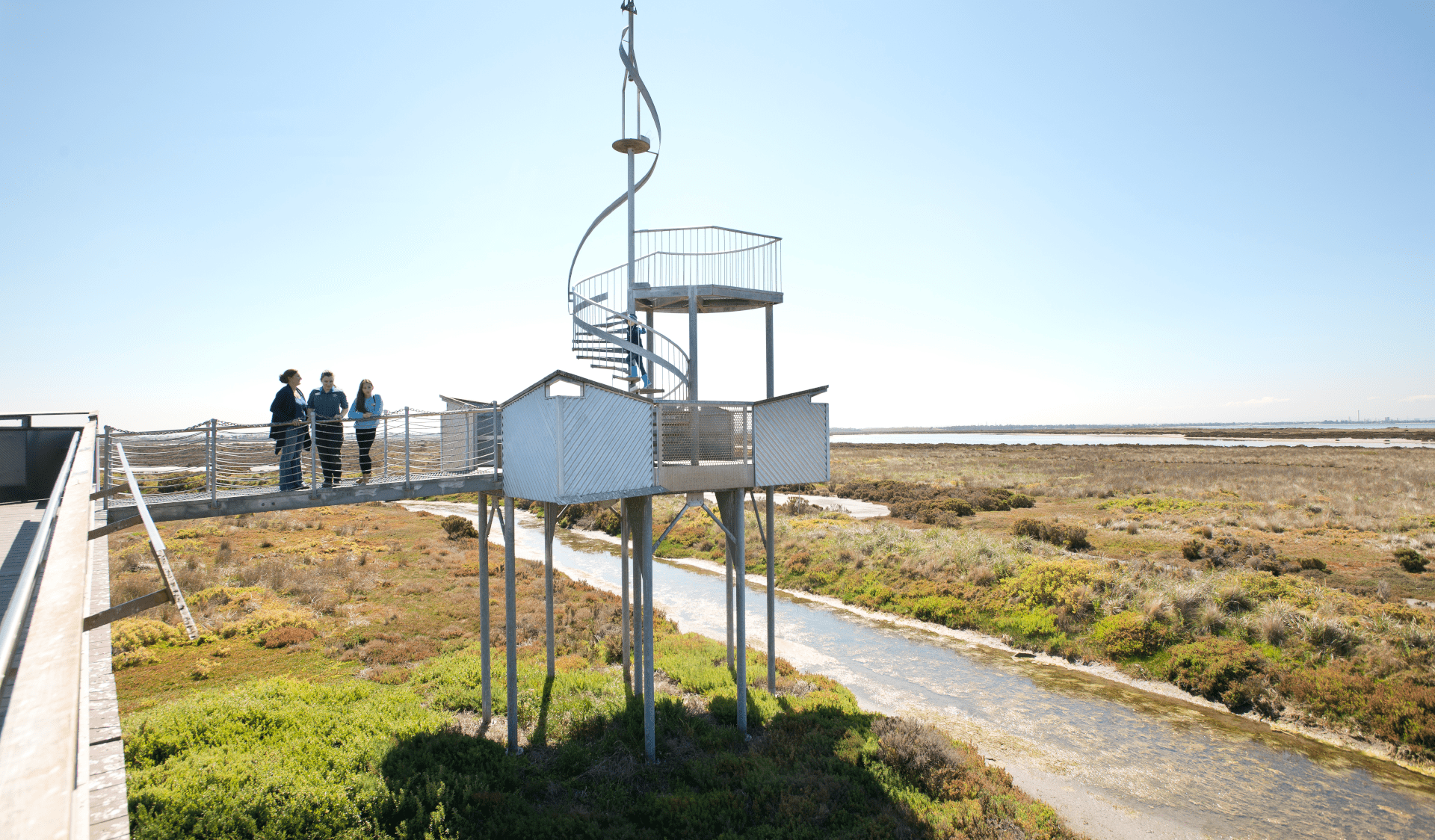 Three people stand on the walk bridge of the Tower at Point Cook Coastal Park