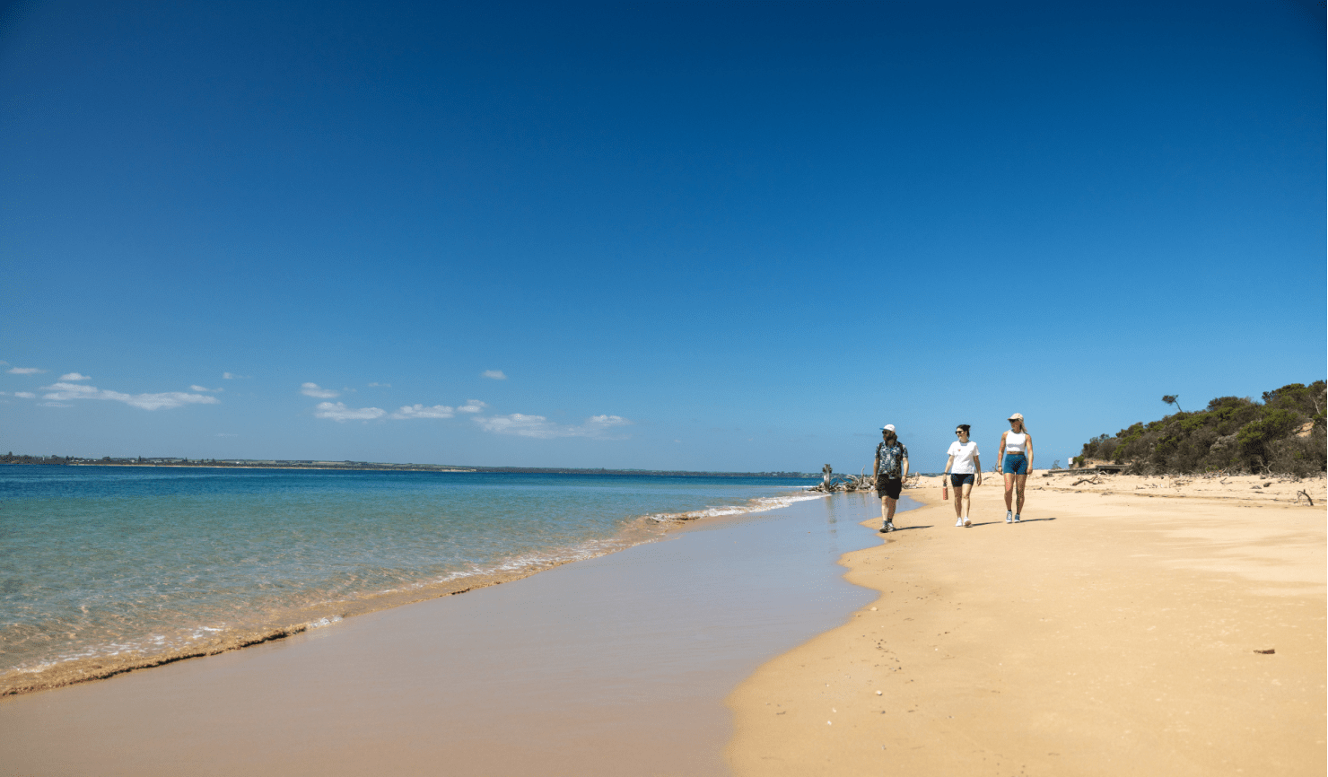 Three people walking along sand near coastline.