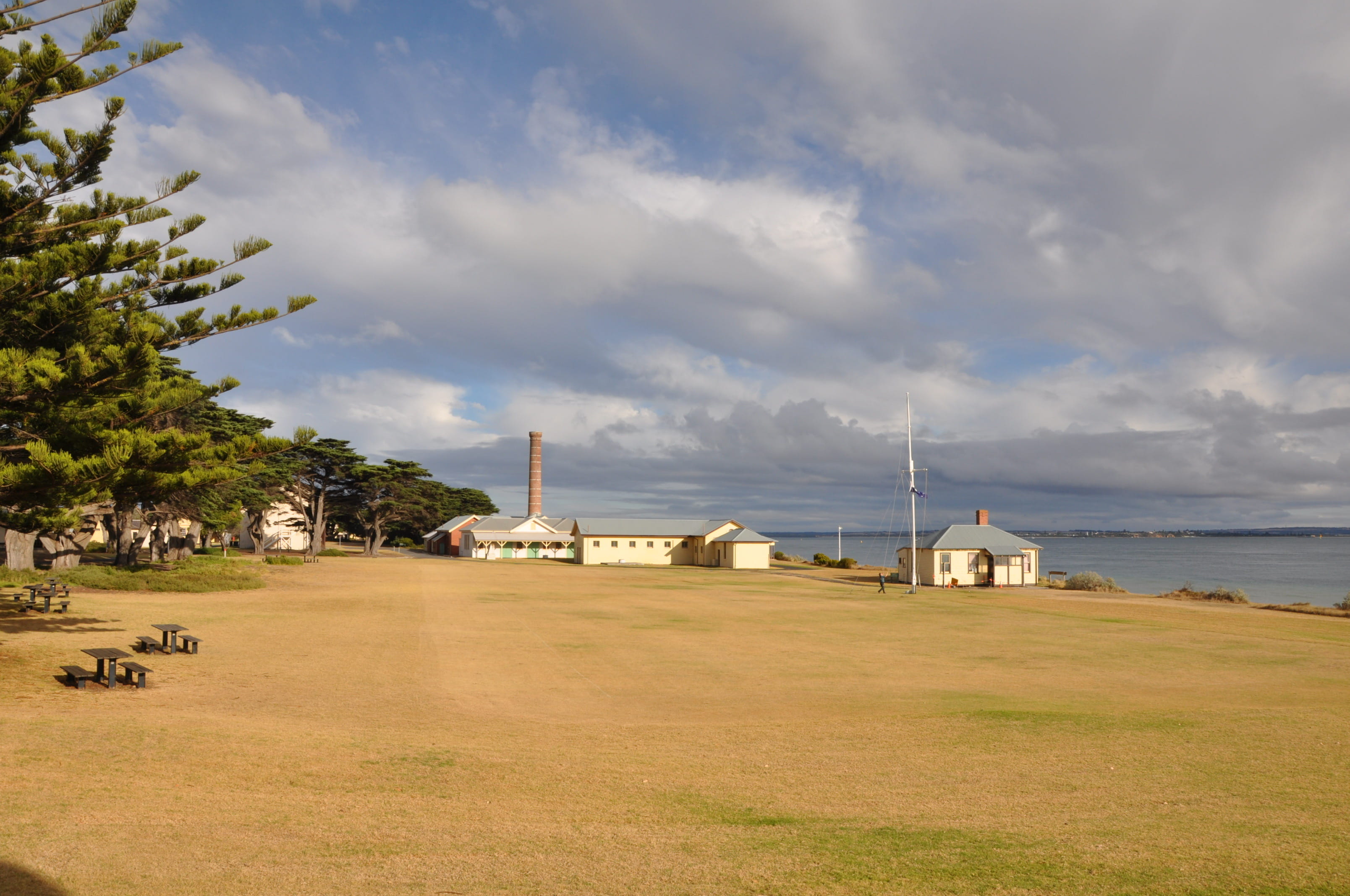 The Quarantine Station at Point Nepean National Park at sunrise. A large stretch of dried grass is in the foreground, with white buildings (Disinfecting Complex) in the background. Grey clouds are in the sky. 