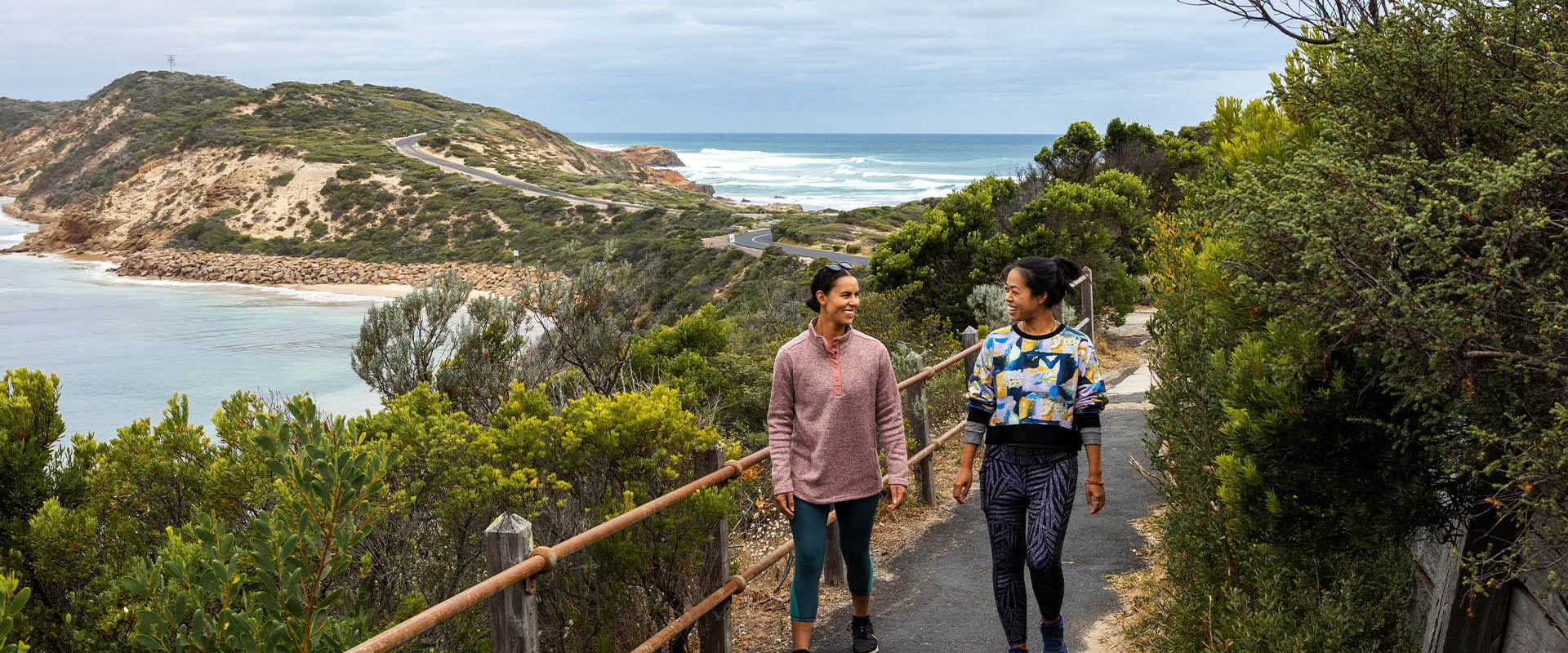Two women walk along a walking path on a narrow peninsula of land in front of the wave-filled ocean and next to the calmer bay of water. 