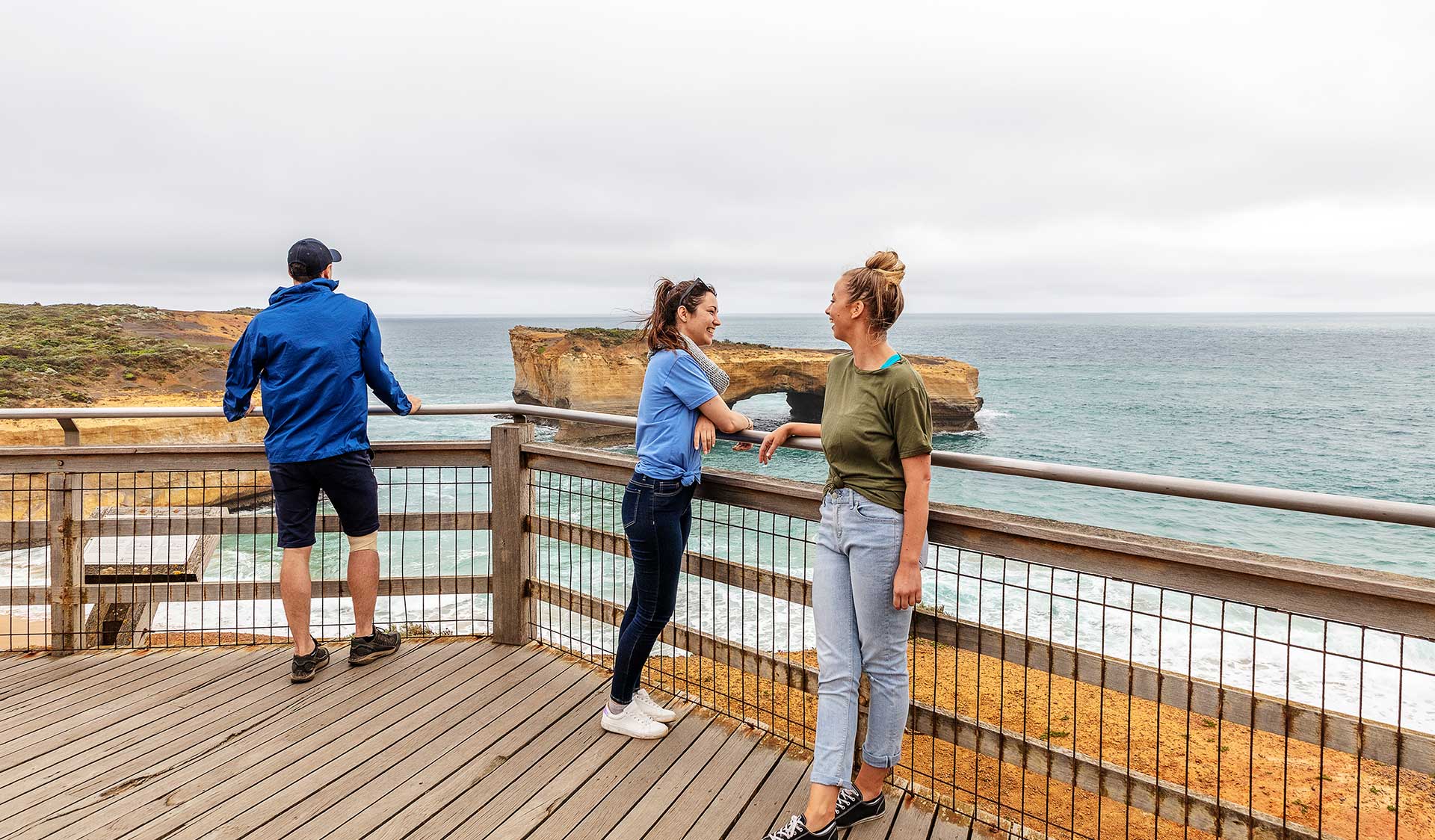 Two women in chat at lookout while a man takes in the view of London Bridge in the Port Campbell National Park.