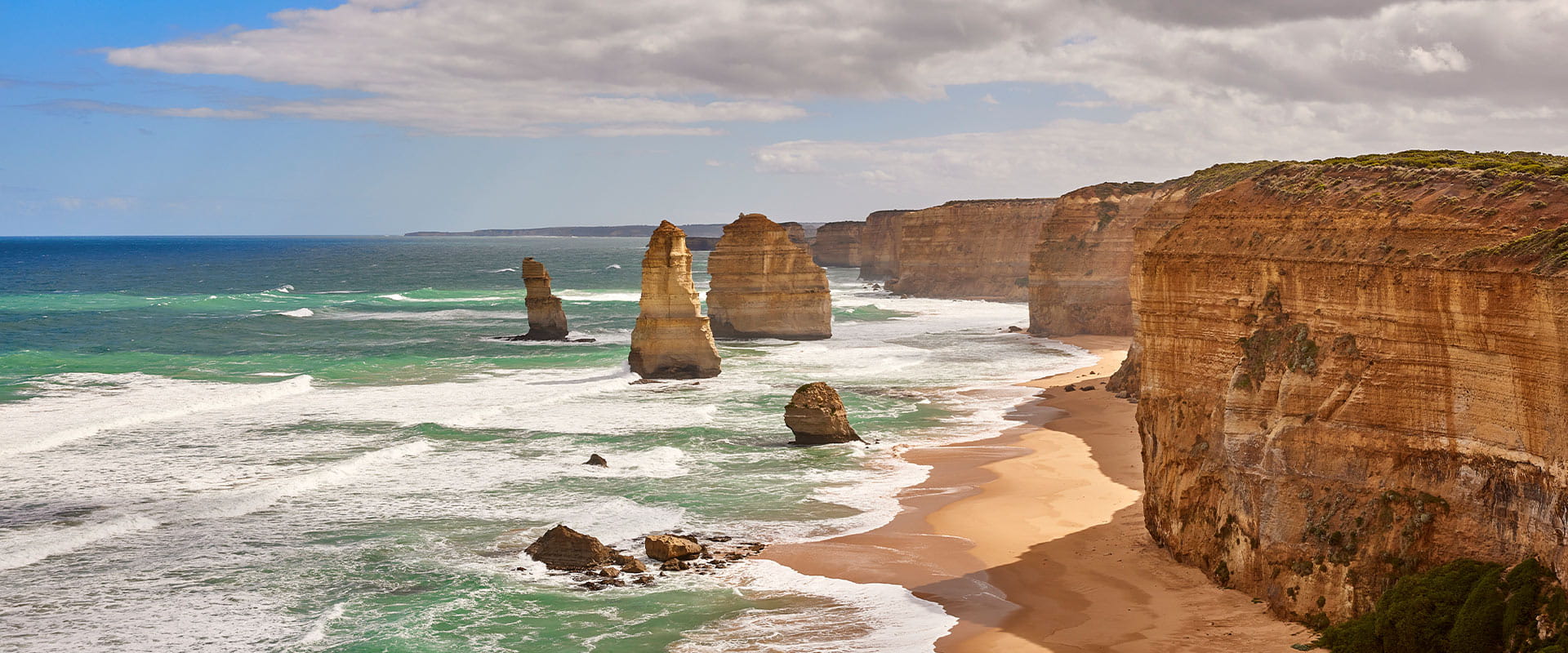 Four large rock stacks stand out of the surf next to tall orange sandstone cliffs above a sandy beach.