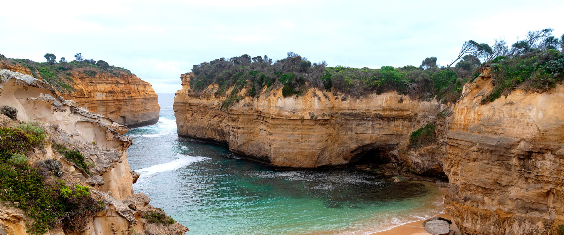 A sandy cove with high green foliage-covered cliffs forming a gorge for the ocean to enter.