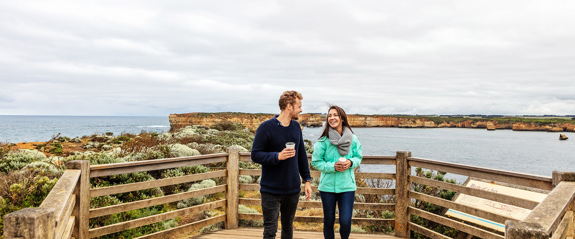 A man and women walk at a lookout overlooking the ocean and sandstone cliffs in the background walk towards the camera.