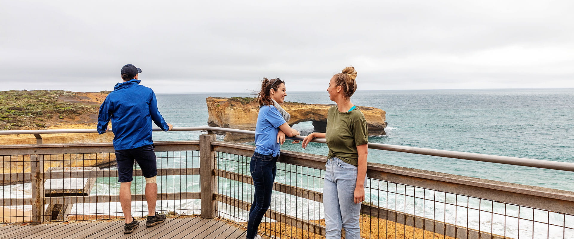 Two women chat at a coastal lookout in front of a sandstone rock stack resembling a bridge.
