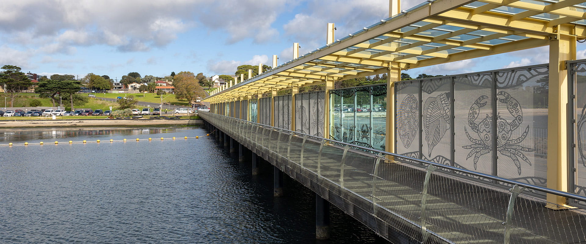 The pier walkway is sheltered and features aboriginal sea life artwork on the glass. Houses and a hill can be seen in the background.