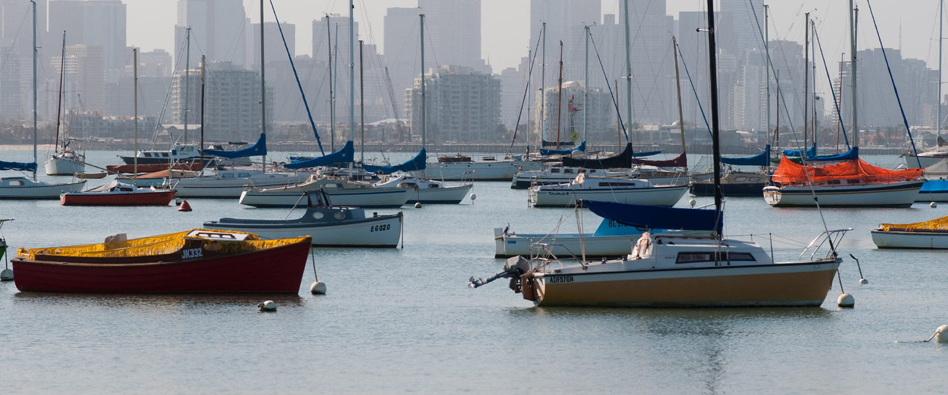 Yachts sit idle, anchored in a protected harbour; the city of Melbourne is in the background, with towering skyscrapers sitting behind the buildings on the foreshore.