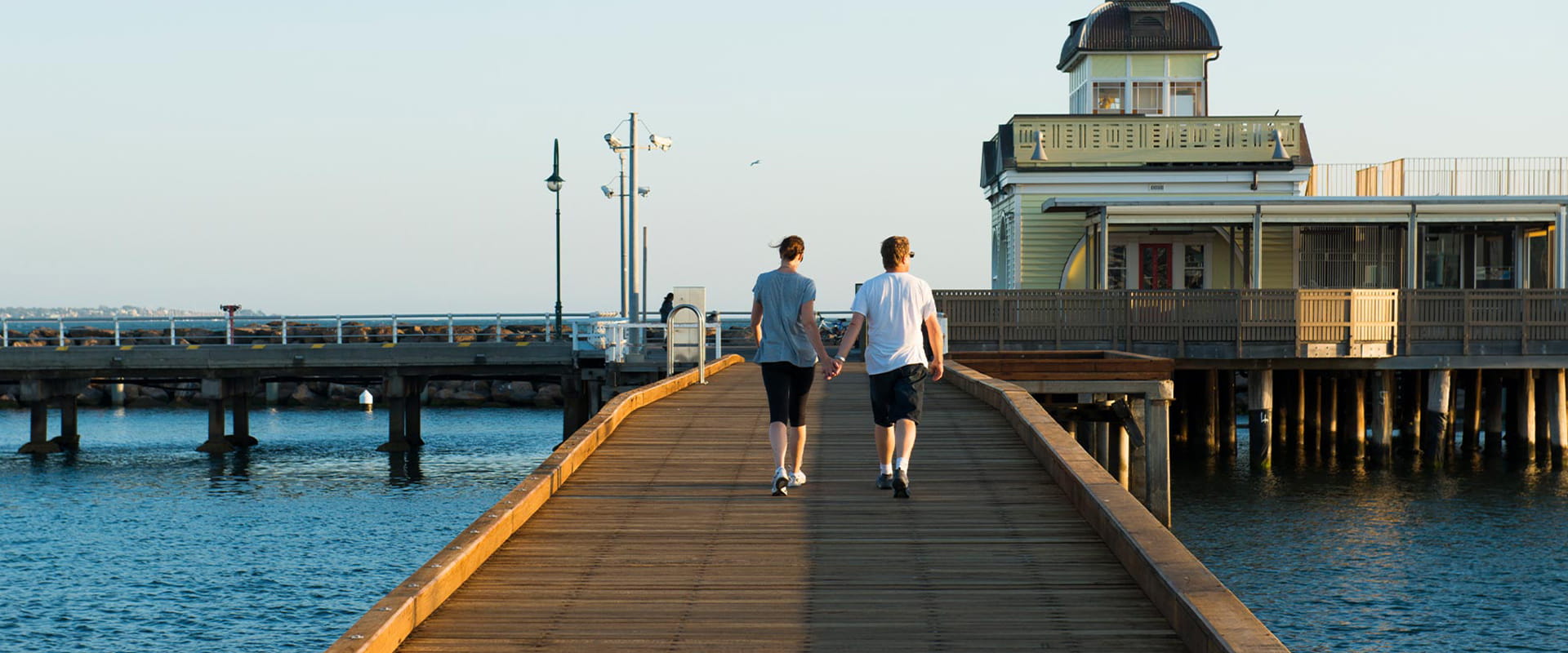 A man and woman hold hands while walking down a wooden pier towards a historic building and a stone breakwater.