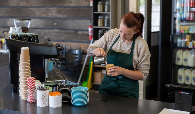 A barista in a green apron pours milk into a cafe latte next to a coffee machine behind the counter.