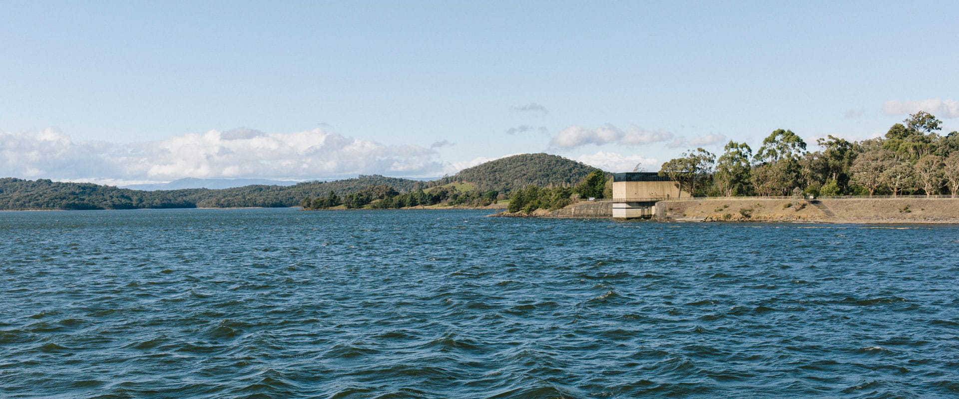 An expansive body of water. Forested hills in the background. At the edge of the lake a concrete walled structure. 