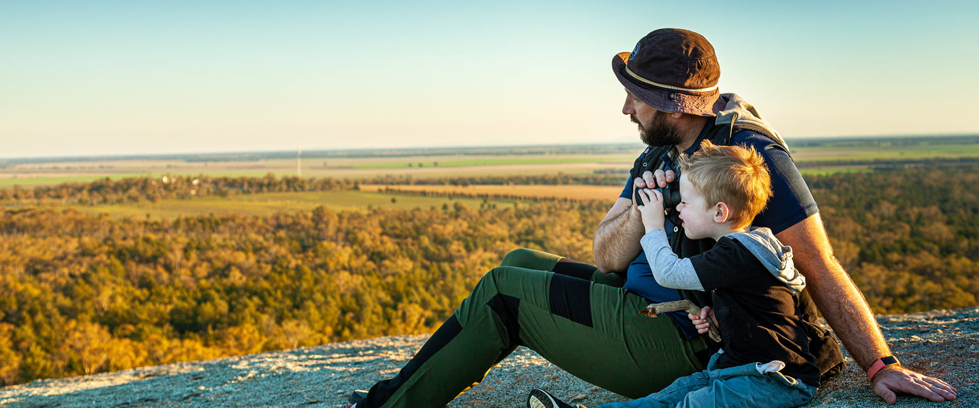 A father and son look through binoculars from a hilltop whilst enjoying the sunset