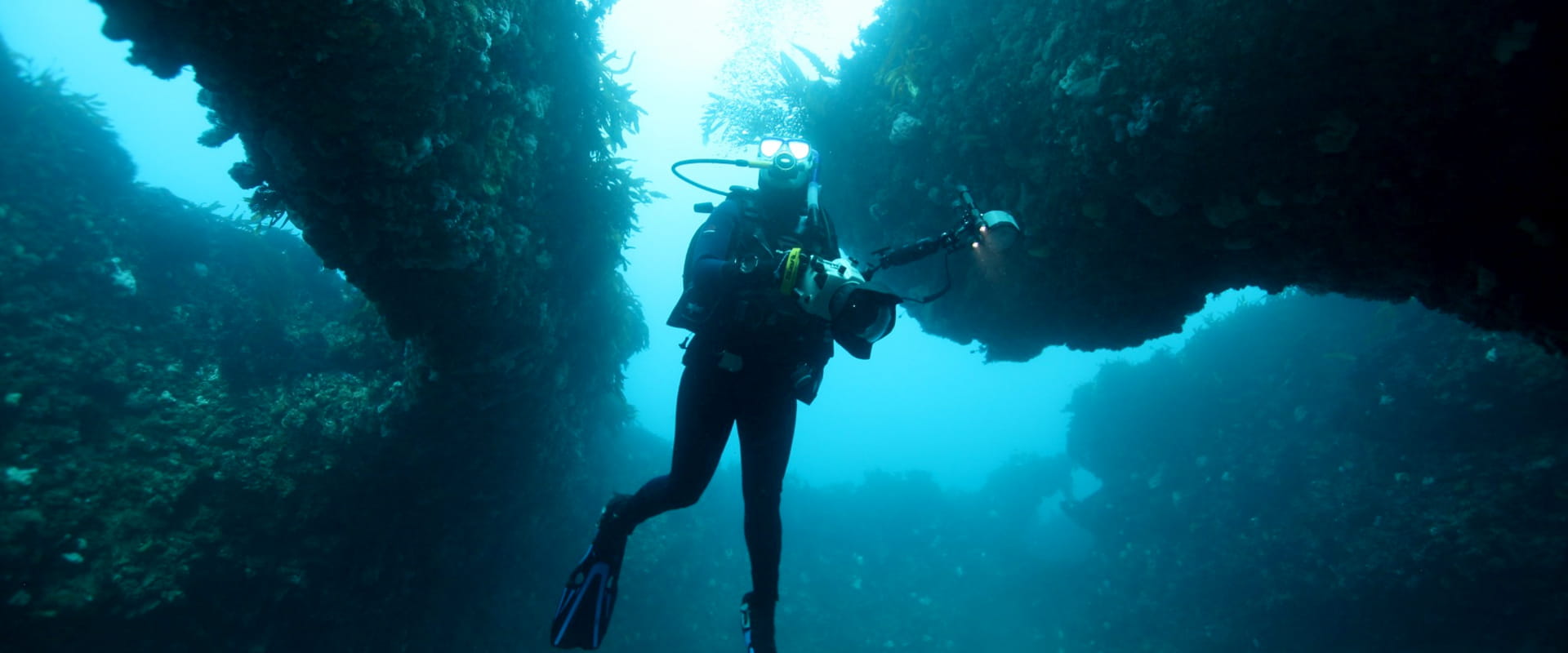A scuba diver is holding a large underwater camera rig and is underwater beneath two rocky arches covered in coral and marine life.