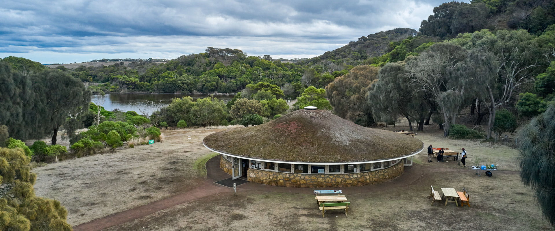 A round mid-century building stands in a beautiful green valley landscape. A body of water in the background. 