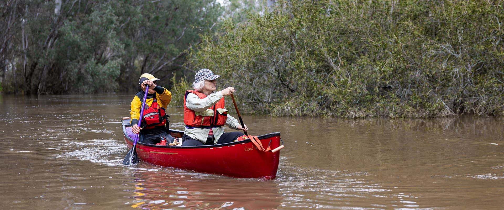 Two people in life-vests and a red canoe rowing along a river with vegetation on the banks in the background.