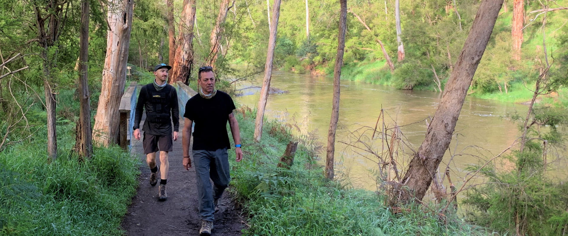 Two men walk alongside a deep green fast flowing river in lush green parkland