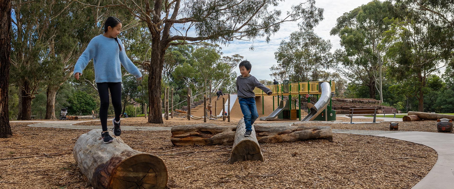 Two kids laughing as they play on logs next to a curving path with a playground in the background.