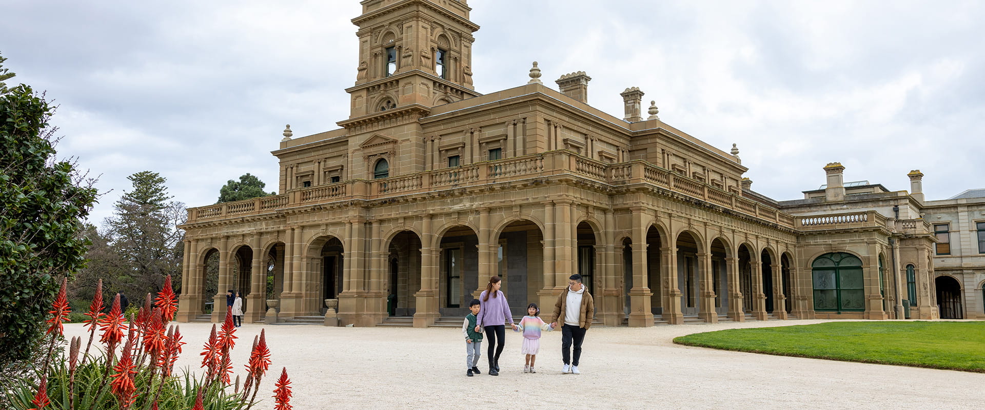 A young Chinese family of four walks hand in hand in front of a sandstone stately home.