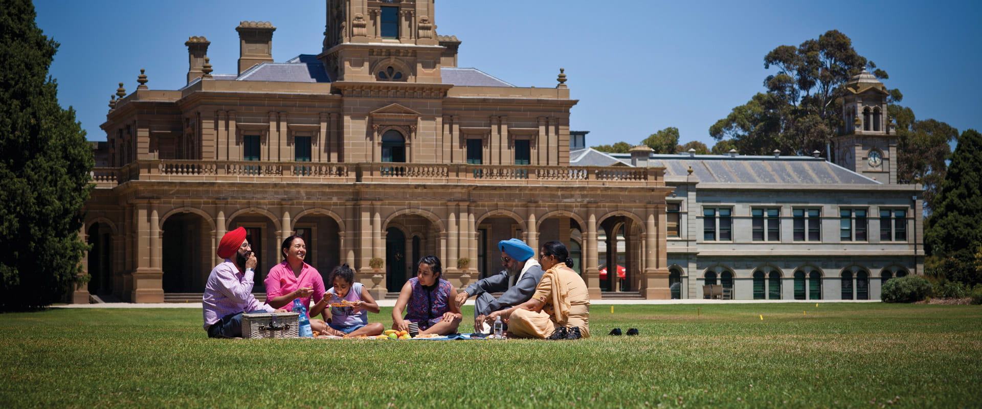 A family of six enjoy a picnic on a green, manicured lawn in front of a multi-storey sandstone mansion featuring striking columns and archways.