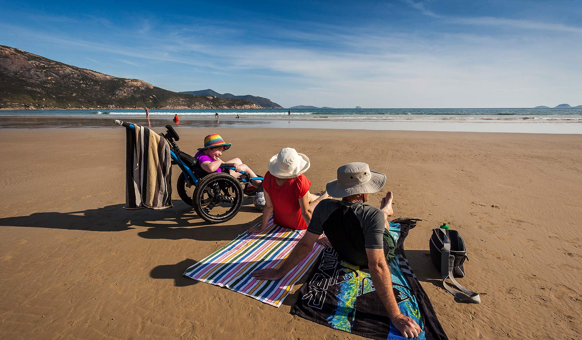 A father and mother sit on Squeaky Beach with their daughter in an all terrain accessibility wheelchair.