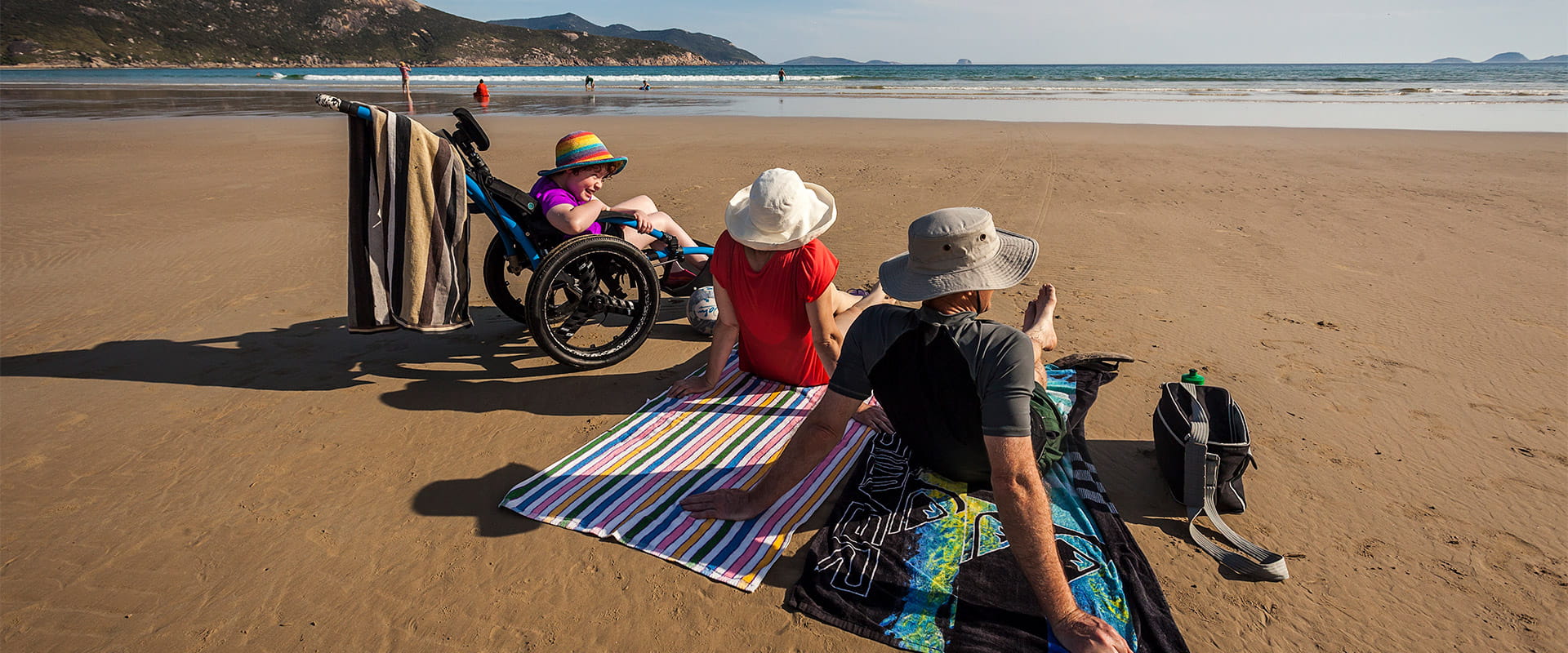 A child wearing a rainbow hat sits in a beach wheelchair while smiling at two adults sitting on beach towels on the sand.
