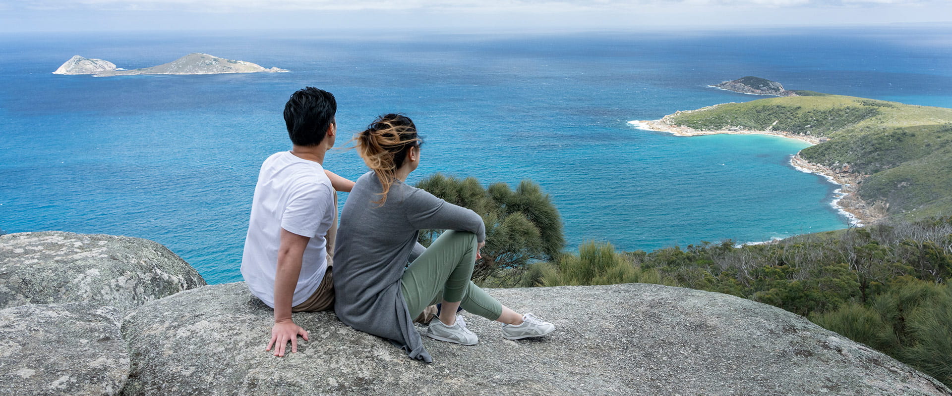 A couple sit on top of some rocks looking at a spectacular ocean view and remote coastal beaches