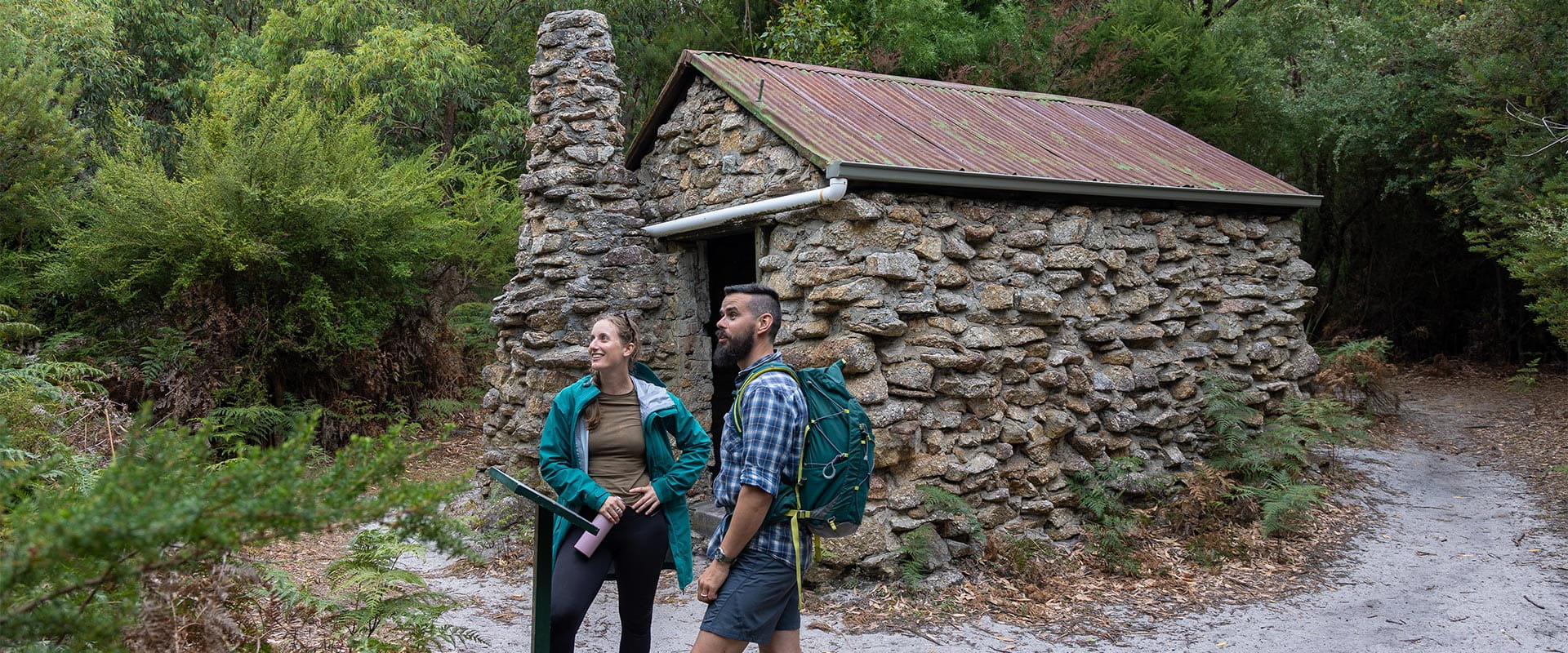 A woman and man wearing hiking gear looking at the view while standing in front of an information board with a hut in the background.