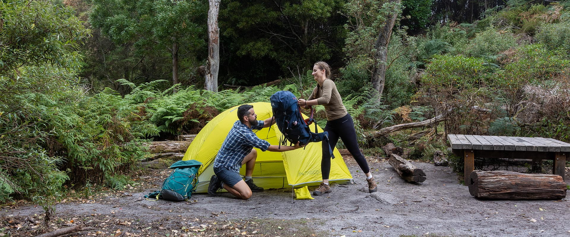 A kneeling man reaching out to take a hiking pack from a standing woman outside a yellow tent, surrounded by vegetation.