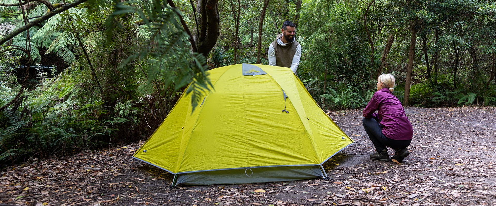 A man and woman finish setting up a yellow tent surrounded by ferns and trees in the background.