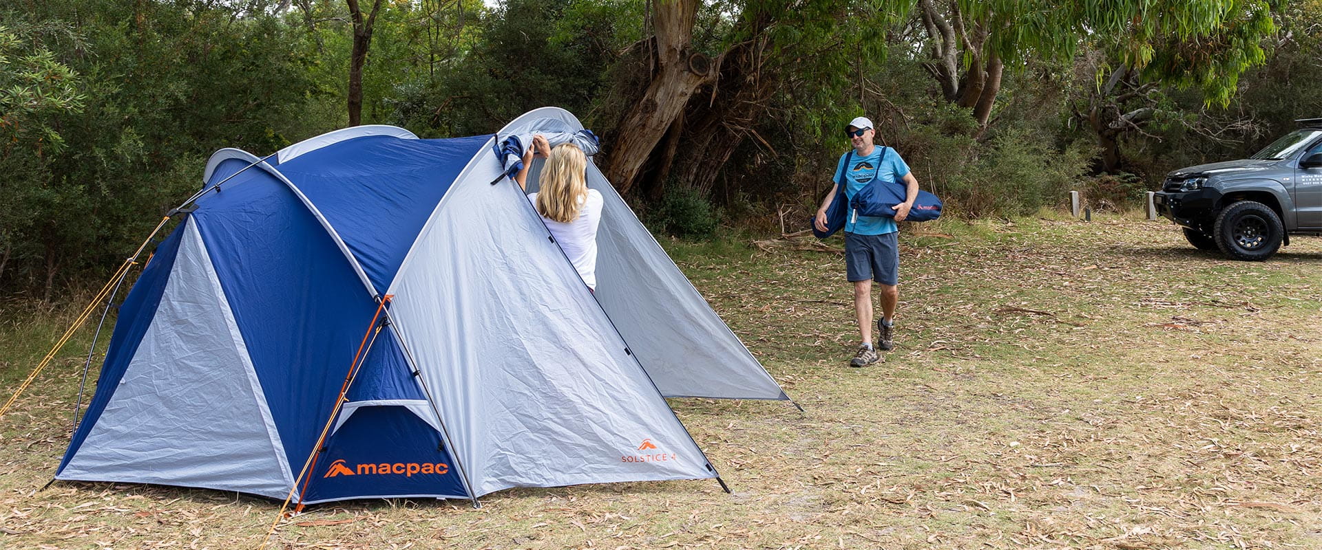 A woman rolls up a tent flap as a man moves more camping gear from the car to the tent.