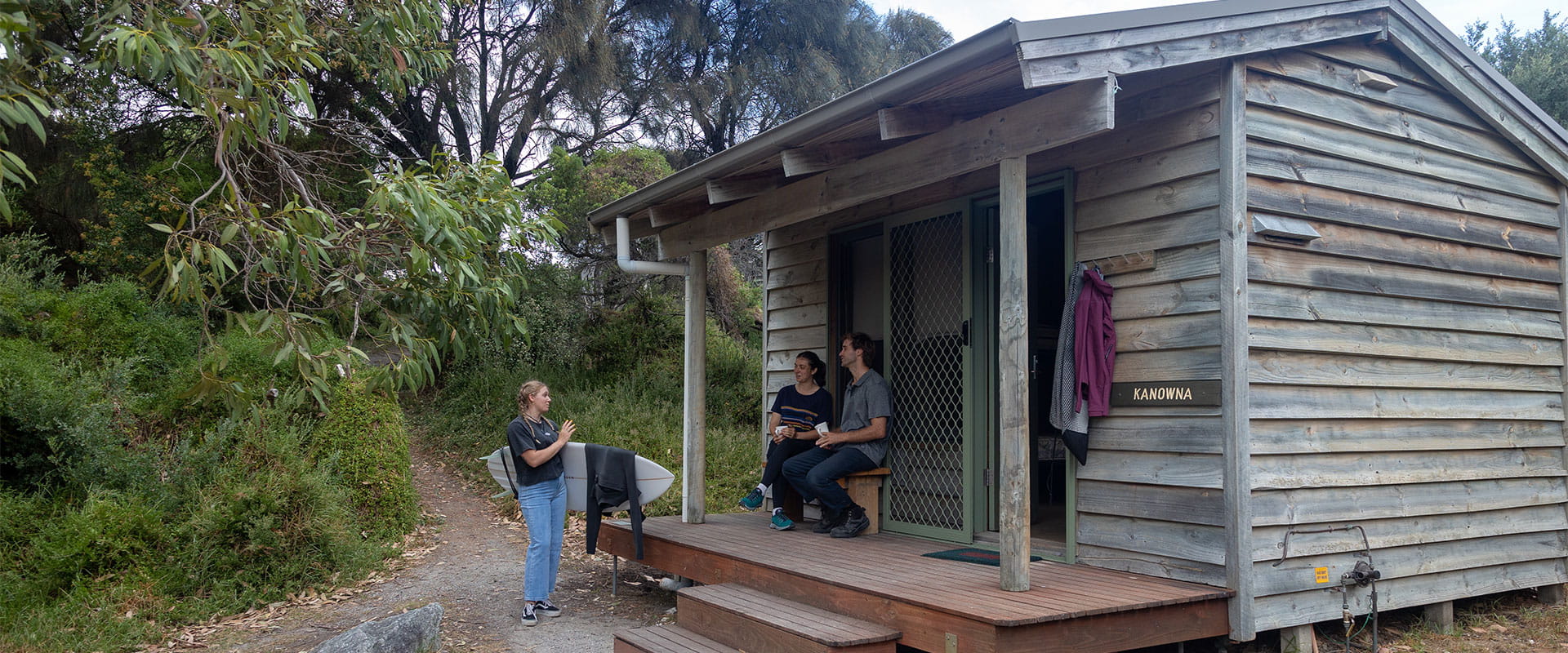 A woman holding a surfboard talks to a woman and man holding mugs and sitting outside a wooden hut named Kanowna.