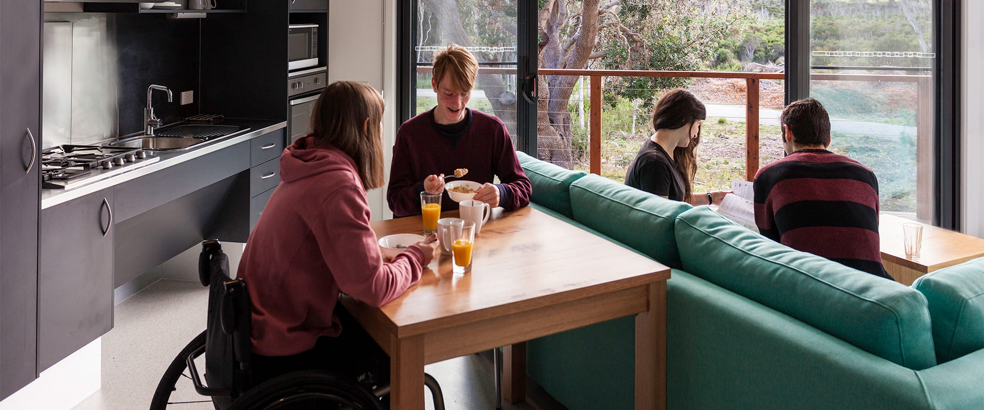 A woman in a wheelchair sits at a wooden table while eating breakfast and talking with a man, while another man and woman sit on a green couch in the background looking at printed information.