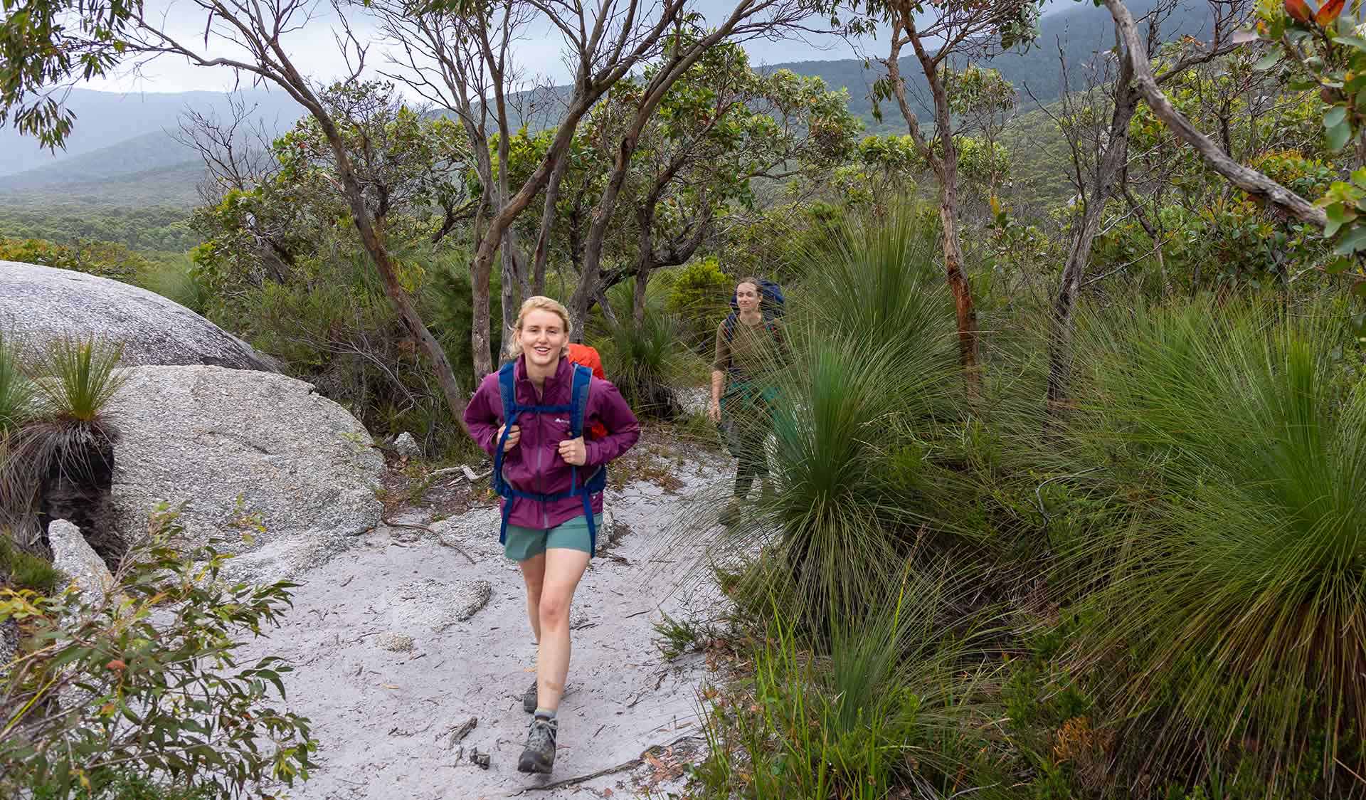 Two women walk along the track between granite rocks and grass trees on the Southern Circuit hiking trail at Wilsons Promontory National Park