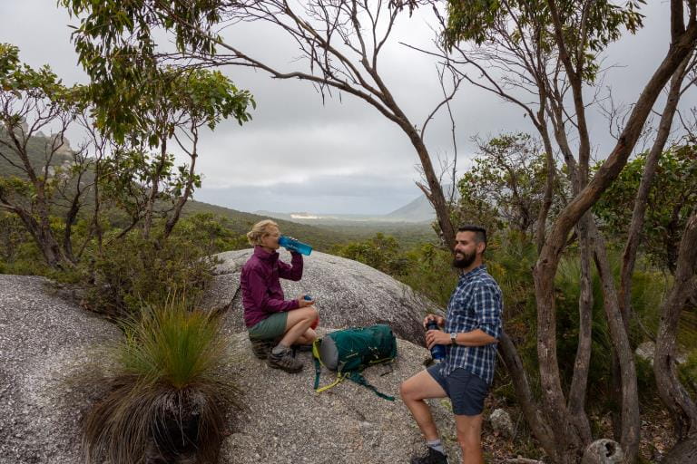 A man and women stop for a drinks break next to granite rocks on the Southern Circuit hiking trail at Wilsons Promontory National Park