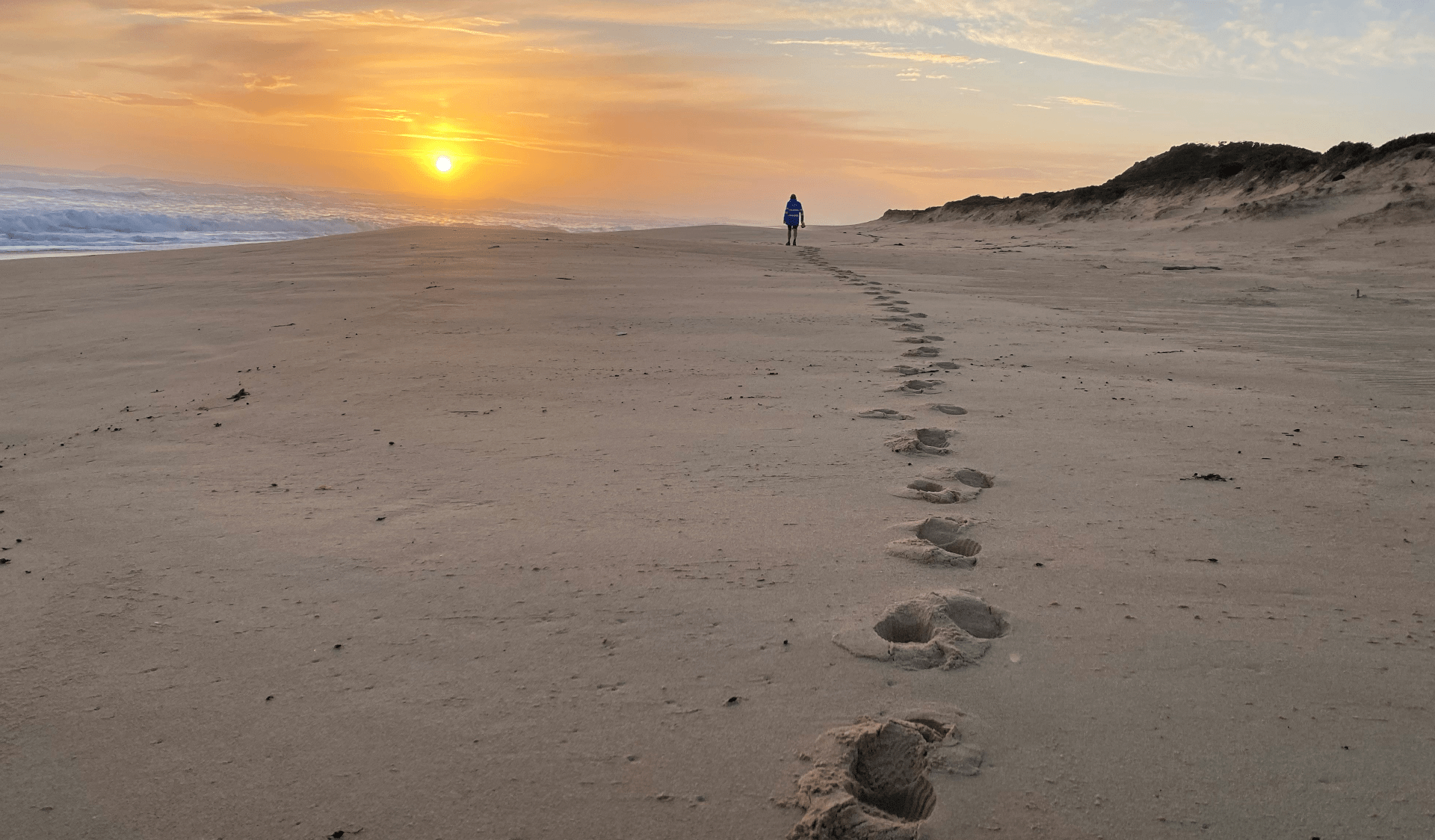 A man walks along Willimsons Beach with footprints behind at sunset