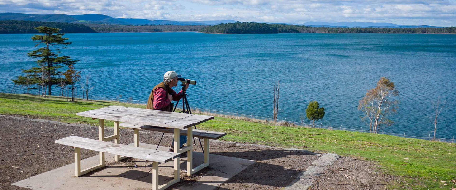 A man sits on a picnic table with a binocular looking at the wildlife across the reservoir.