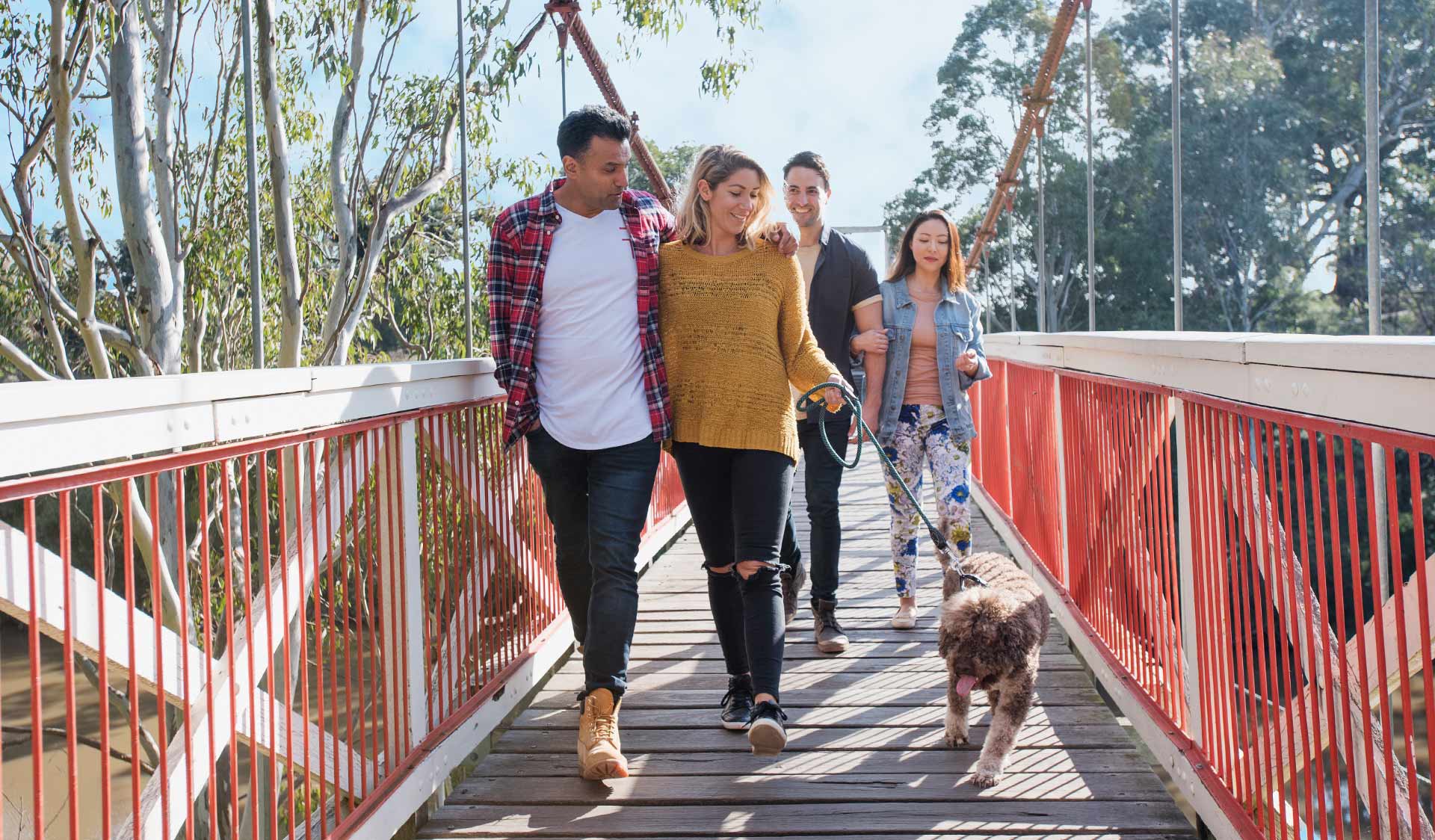 A young couple walking their dog across a bridge with two friends following behind