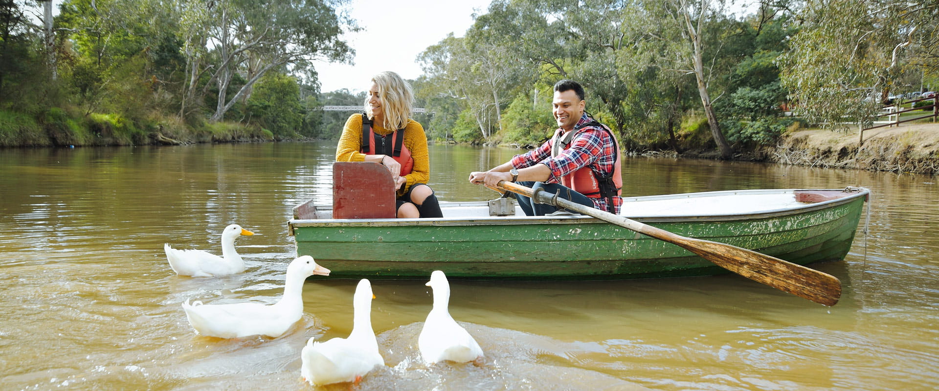 A woman and man wearing lifejackets sit in a small rowboat as four white ducks paddle around them in a river, with trees stretching out from the banks on either side.