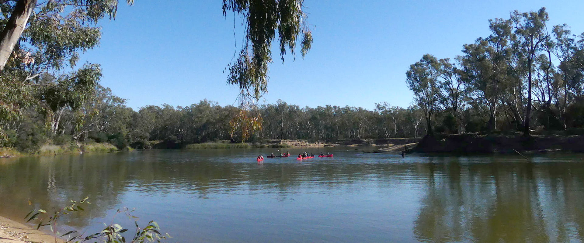 A group of people in red Kayaks gather in the centre of a lake surrounded by rugged bushland.